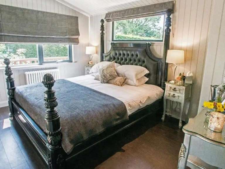 Ein großes Bett mit einem dunklen Kopfteil und einem grauen Überwurf steht in einem Zimmer mit weißen Wänden und großen Fenstern.