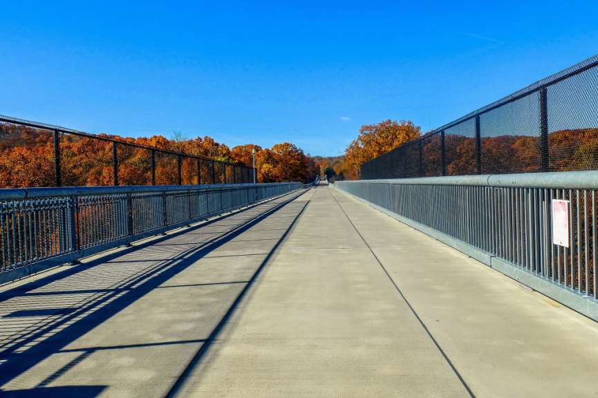 Eine breite Betonbrücke führt auf einen bewaldeten Hügel zu, der von einer klaren blauen Himmel überragt wird.