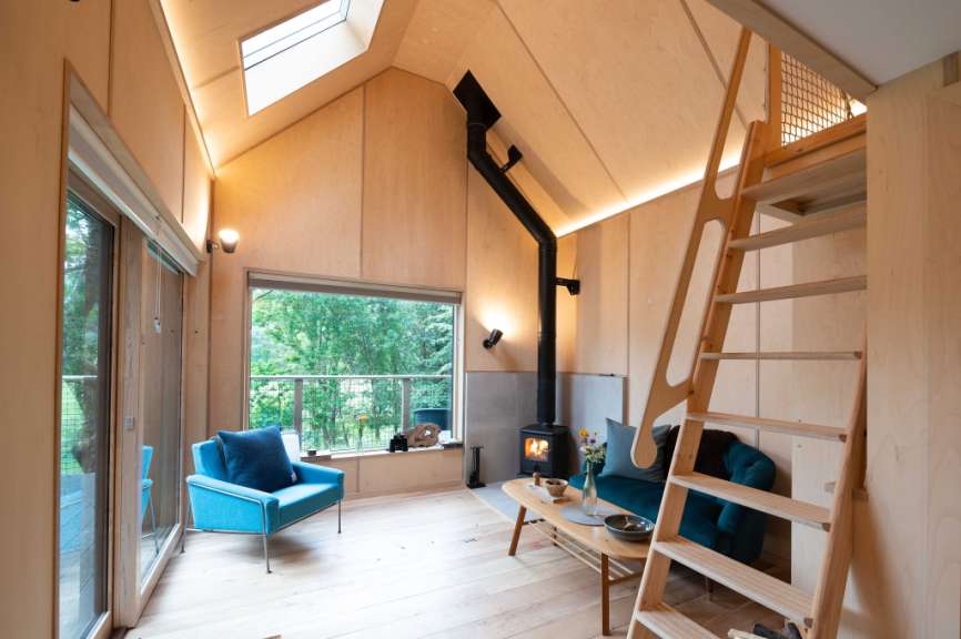 Ein gemütliches holzverkleidetes Wohnzimmer mit einem Kaminofen, einem blauen Sessel und einer Leiter, die zu einem Hochbett führt.