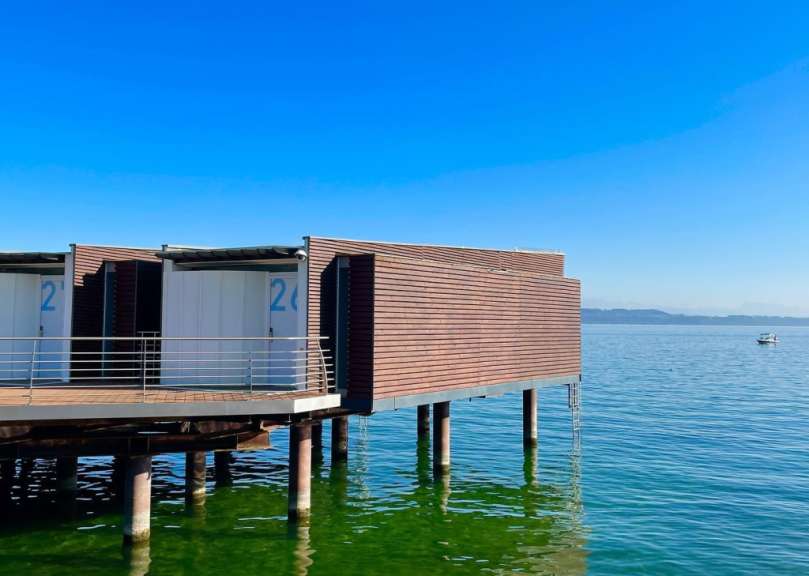 Holzhütten auf Stelzen im klaren Wasser unter blauem Himmel mit einem Boot am Horizont.