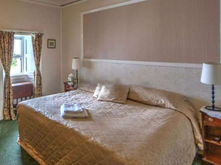 Ein Schlafzimmer mit einem großen Bett, Nachttischen und einem Fenster mit Vorhängen wird gezeigt.