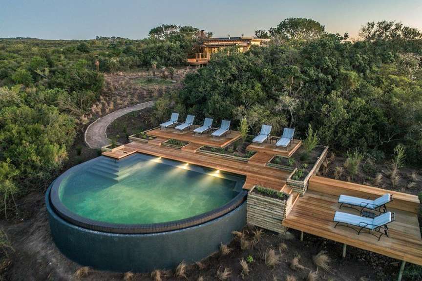 Ein runder Infinity-Pool mit einer Holzterrasse, Sonnenliegen und einem Haus in einer bewaldeten Landschaft.