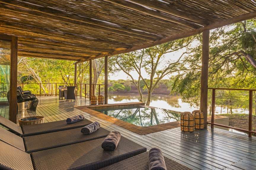 Ein überdachter Holzdeck mit Liegestühlen und einem Pool blickt auf einen ruhigen Fluss und üppige Vegetation.