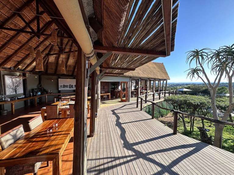 Eine offene, mit Holz verkleidete Veranda mit Essbereichen und Blick auf eine Landschaft mit Bäumen und dem Meer unter einem strahlend blauen Himmel.
