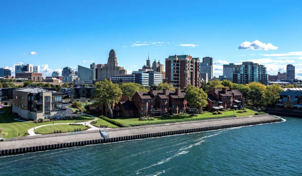 Eine breite Wasserstraße mit einem Ufer, an dem sich ein Komplex von roten Ziegelhäusern und modernen Wohngebäuden befindet, erstreckt sich im Vordergrund, während im Hintergrund eine Stadt Skyline unter einem blauen Himmel mit einigen Wolken aufragt.