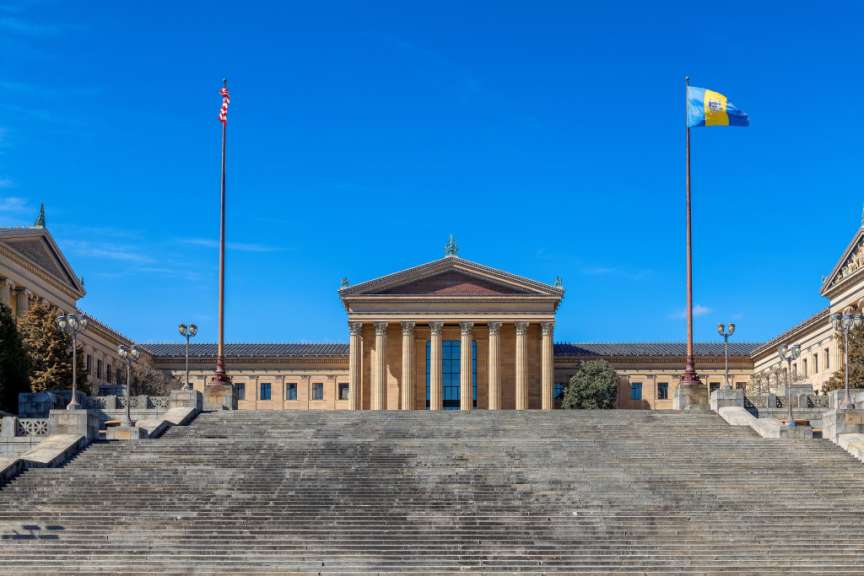 Ein breites Freitreppenensemble führt zu einem hellgelben Gebäude mit Säulenvordach unter einem strahlend blauen Himmel mit zwei Fahnenmasten.