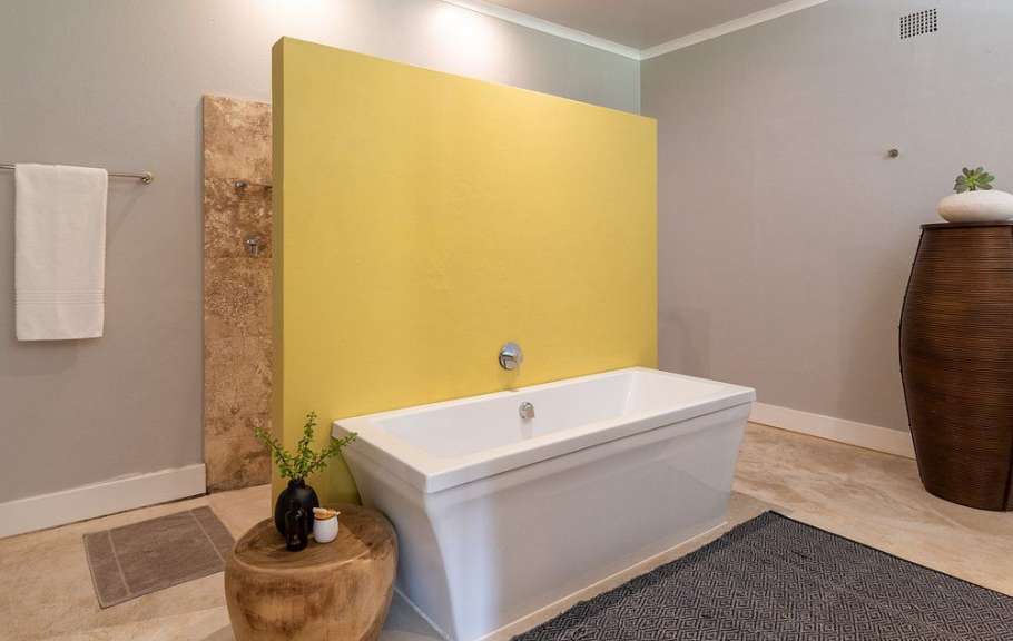 In einem Badezimmer steht eine weiße Badewanne vor einer gelben Wand, daneben ein Holzhocker mit Dekoration.