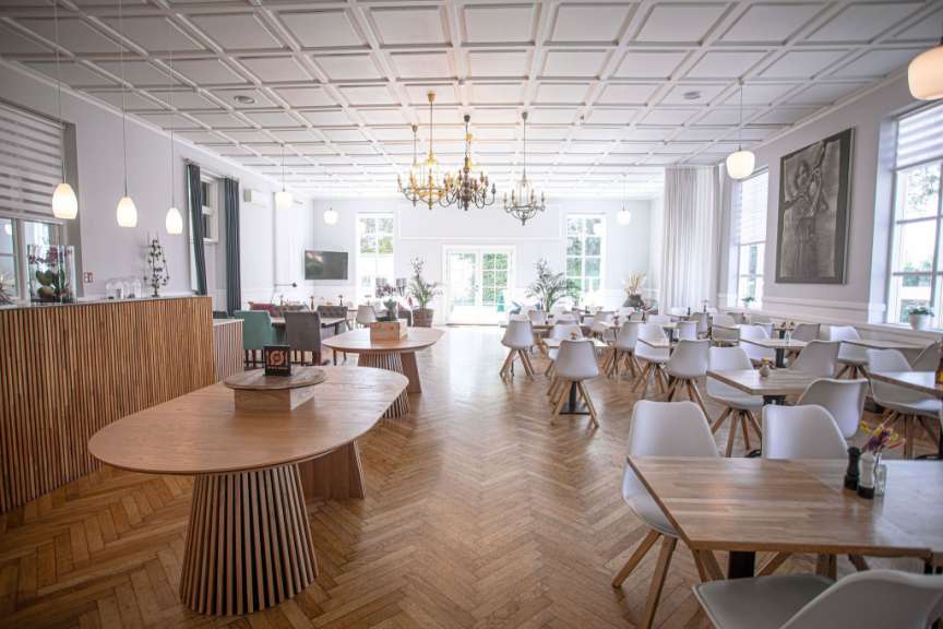 Ein heller und geräumiger Speisesaal mit vielen Holztischen und weißen Stühlen ist mit einer Kassetten decke und Kronleuchtern ausgestattet.