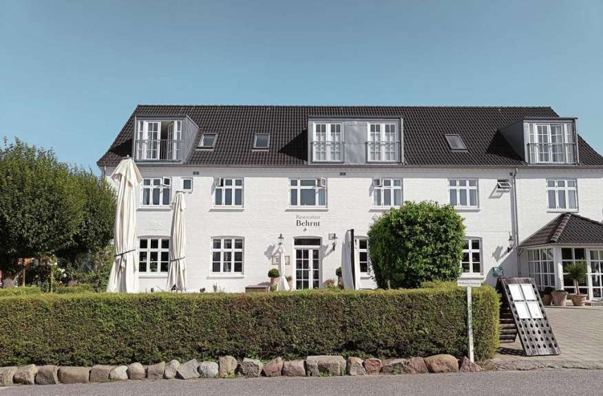 Ein weißes Gebäude mit dunklem Dach und zahlreichen Fenstern, das durch eine Hecke und zwei Sonnenschirme verdeckt ist.