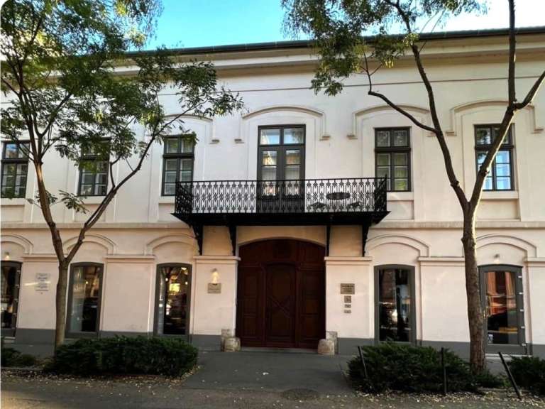 Das Bild zeigt die Fassade eines weißen Gebäudes mit einem Balkon, großen Holztüren und mehreren Fenstern, umgeben von Bäumen und Büschen.