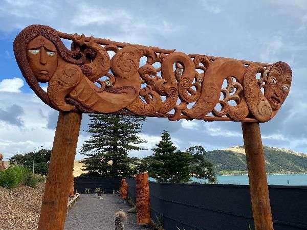 Ein geschnitztes Holzportal mit traditionellen Māori-Motiven steht vor einer Wasserlandschaft unter einem bewölkten Himmel.