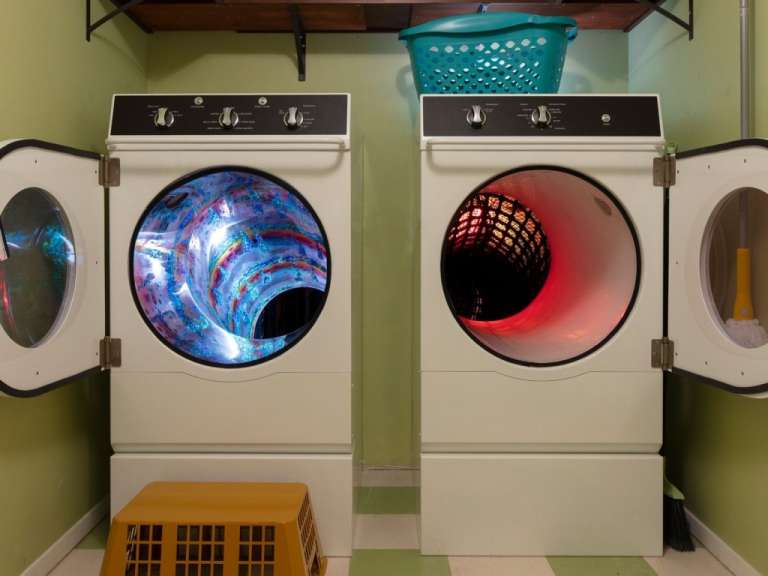 Ein Paar Waschmaschinen stehen nebeneinander, ihre Türen sind geöffnet und enthüllen ihre Trommeln, die mit bunten Mustern gefüllt sind, während daneben ein Mop und ein Besen stehen.