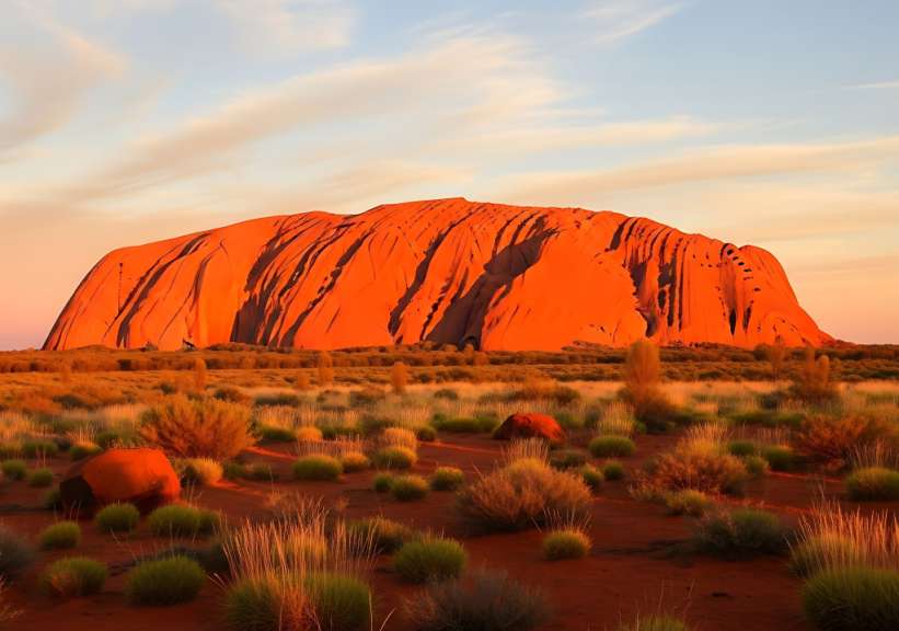 Ein riesiger orangeroter Monolith erstreckt sich am Horizont unter einem hellblauen Himmel mit zerstreuten Wolken und ist von trockener Vegetation und rotem Sand umgeben.