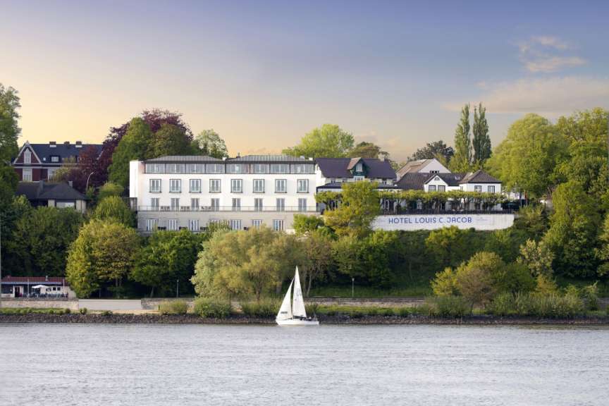 Das Bild zeigt ein Hotel mit dem Namen "Hotel Louis C. Jacob" am Ufer eines Flusses mit einem Segelboot im Vordergrund.