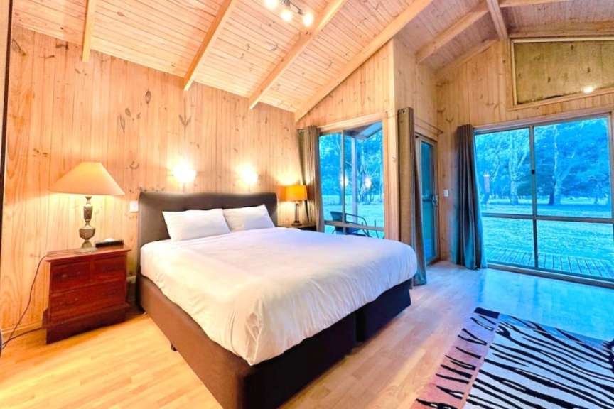 Das Bild zeigt ein Schlafzimmer mit Holzwänden und -decke, einem großen Bett und bodenhohen Fenstern, die auf eine bewaldete Landschaft blicken.