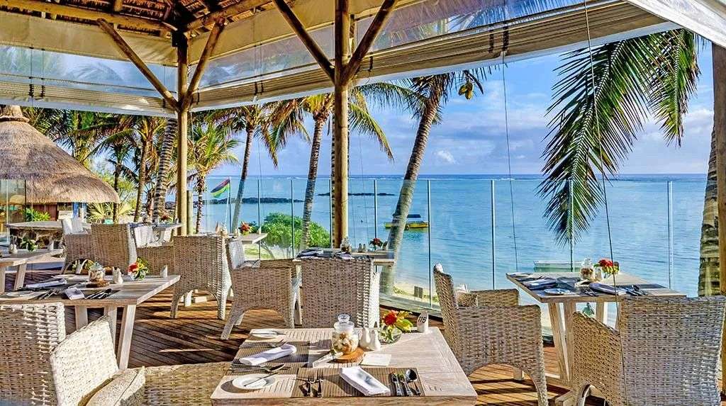 Ein Tisch ist in einem Restaurant mit Blick auf das Meer gedeckt, das von Palmen und einem tropischen Himmel umrahmt wird.