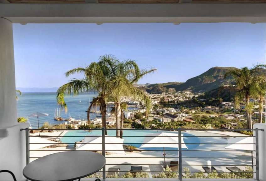 Blick von einem Balkon auf ein Meer mit Booten, einen Pool mit Sonnenschirmen und eine Stadt am Hang unter einem bergigen Horizont.