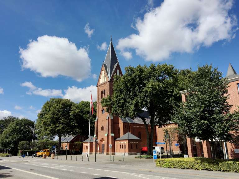 Das Bild zeigt eine rote Backsteinkirche mit einem spitzen Turm, umgeben von Bäumen und im Hintergrund einer Straße mit Fahrzeugen an einem sonnigen Tag mit Wolken.