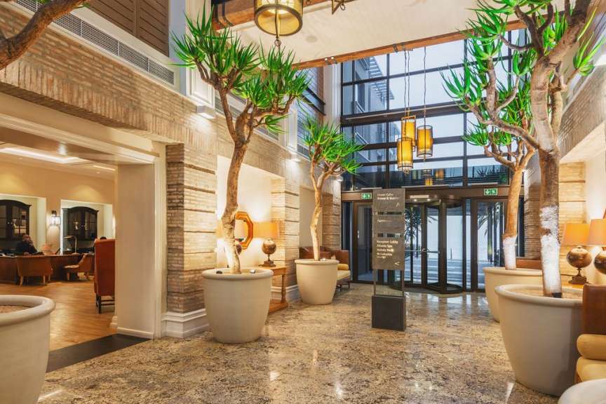 In einer gut beleuchteten Hotellobby mit hohen Pflanzen, die von großen, weißen Töpfen umgeben sind, stehen mehrere orangefarbene Sessel und eine Rezeptionstheke in einem separaten Bereich.
