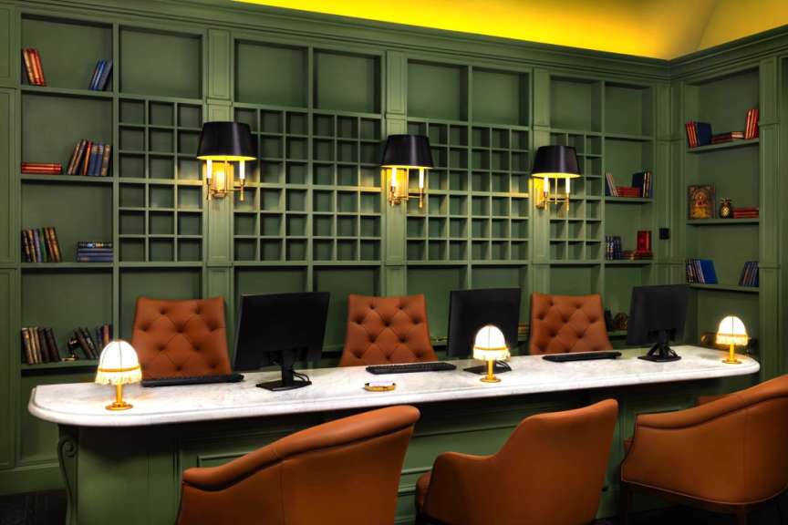 Ein Schreibtisch mit vier Computern, der von braunen Ledersesseln umgeben ist, steht vor einem grünen Bücherregal.