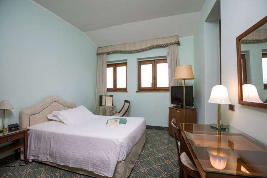 Ein Hotelzimmer mit einem Bett, zwei Fenstern, einem Schreibtisch mit Lampe und einem Spiegel wird gezeigt.