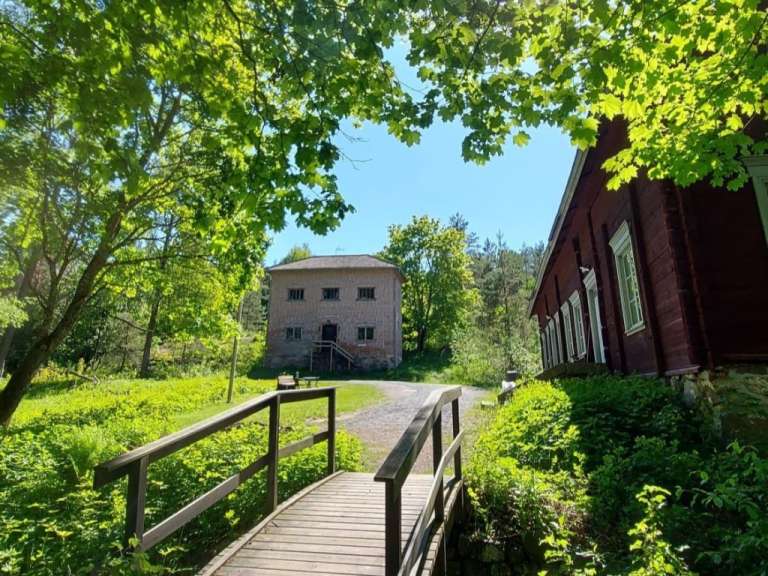 Ein hölzerner Steg führt zu einem Backsteingebäude, während rechts ein rotes Holzhaus zu sehen ist, umgeben von üppigem Grün unter klarem Himmel.