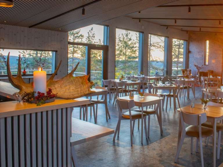 Ein Restaurant mit großen Fenstern und Blick auf eine Waldlandschaft, dekoriert mit einem Rentiergeweih und einer Kerze.