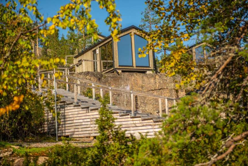 Ein modernes Holzhaus mit großen Fenstern ist auf einem felsigen Hang gebaut, der über eine Holztreppe mit Geländer zugänglich ist und von Bäumen mit gelben und grünen Blättern umgeben ist.
