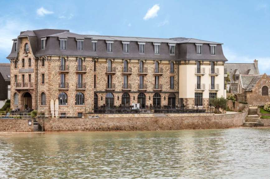 Ein imposantes Steingebäude mit mehreren Etagen und einem Mansarddach steht am Ufer eines Gewässers, flankiert von kleineren Nebengebäuden und umgeben von einer stoneigen Ufermauer.