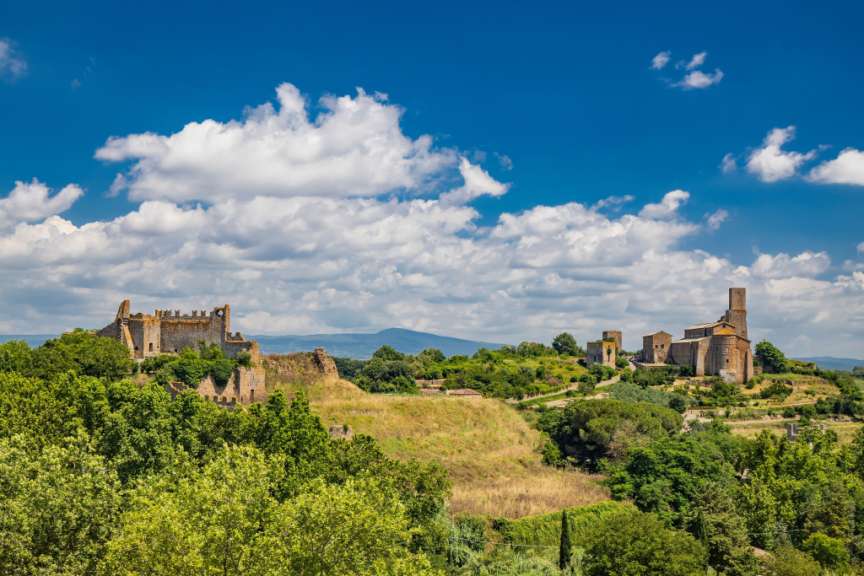 Auf einem Hügel mit viel Grün und blauen Himmel mit Wolken befinden sich Ruinen und Gebäude, die eine alte Stadt darstellen.
