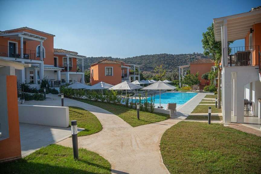 Ein Resort mit pastellfarbenen Gebäuden, einem Pool und gepflegten Rasenflächen liegt vor einem Hügel unter klarem blauem Himmel.