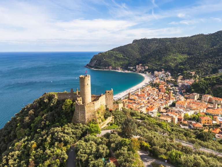 Ein altes Schloss überragt eine Küstenstadt mit einem Strand und dem Meer im Hintergrund unter einem blauen Himmel mit Wolken.