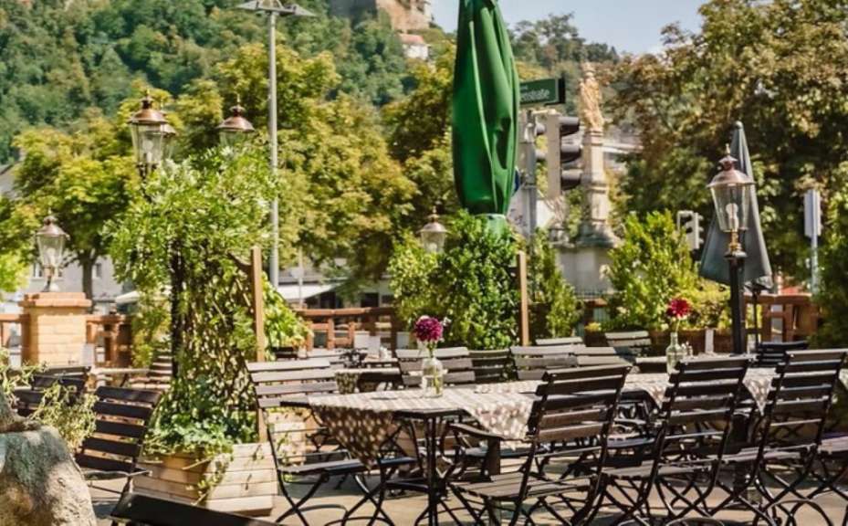 Ein Biergarten mit Holztischen und Stühlen ist im Sonnenschein zu sehen.