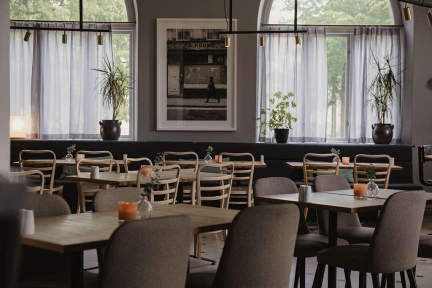 Das Bild zeigt den Innenraum eines Restaurants mit vielen Holztischen und Stühlen, dekoriert mit Kerzen und kleinen Vasen mit Grünzeug.