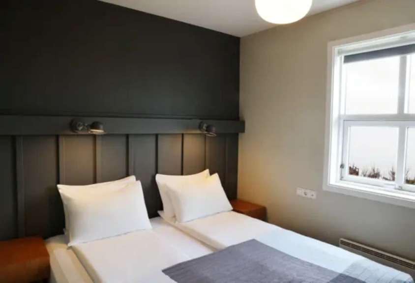 Zwei Betten mit weißen Kissen und einer grauen Decke stehen in einem Zimmer mit dunklem Kopfteil und einem Fenster.