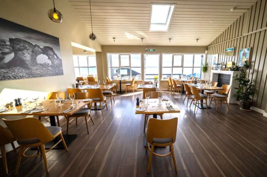 Das Bild zeigt den Essbereich eines Restaurants mit vielen Tischen und Stühlen, die für Gäste gedeckt sind, und große Fenster mit Blick auf das Meer.