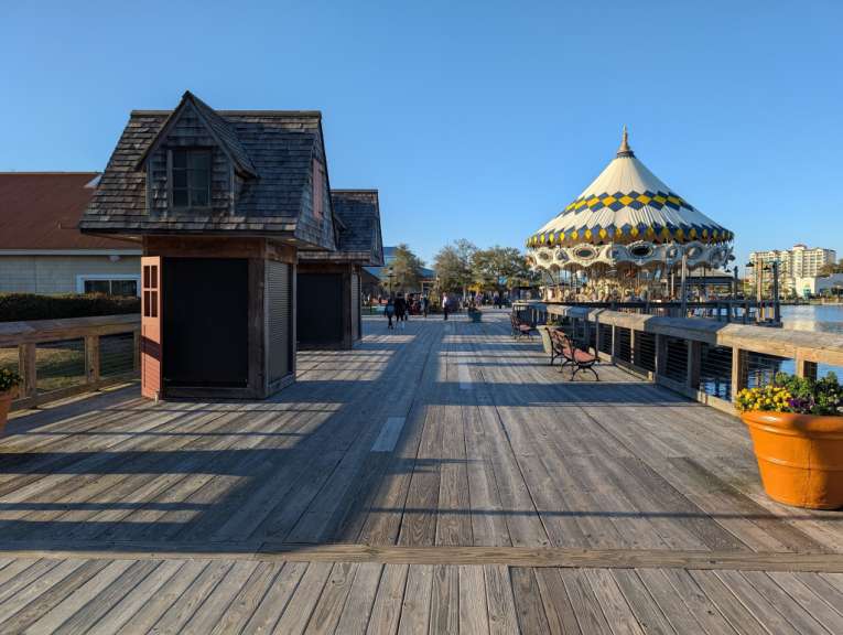 Eine breite Holzpromenade führt zu einem Karussell und mehreren kleinen Holzgebäuden, während im Hintergrund das Meer und ein Hotel zu sehen sind.