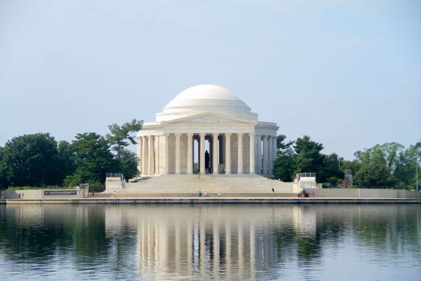Das Bild zeigt das Thomas Jefferson Memorial mit seinen Säulen und der Kuppel, die sich im Wasser des Gezeitenbeckens widerspiegeln, umgeben von Bäumen und mit einigen Besuchern auf den Stufen und am Ufer.