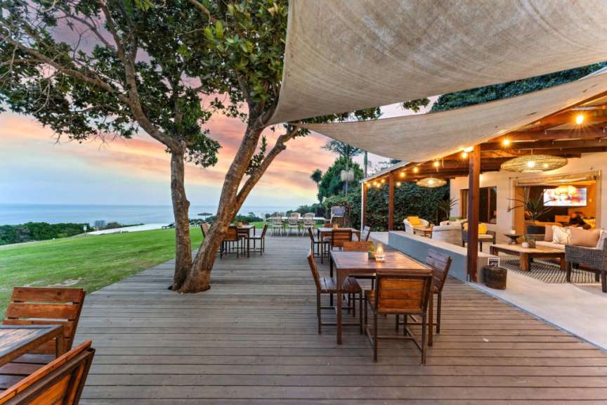 Ein hölzerner Steg führt zu einem Restaurantbereich mit Tischen und Stühlen unter einem Sonnensegel, mit Blick auf einen grünen Hang und das Meer bei Sonnenuntergang.