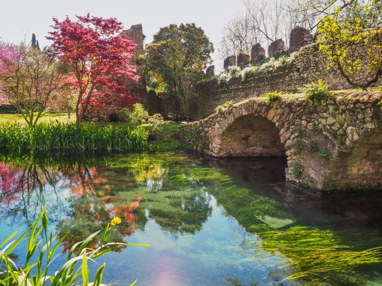 Eine alte Steinbrücke überspannt einen klaren Bach, in dem sich rote Bäume und grüne Pflanzen spiegeln.