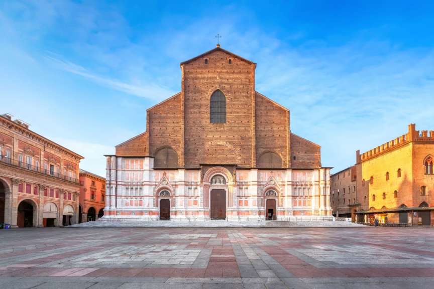 Eine breite Ansicht der Fassade einer Kirche mit einem Platz aus Kopfsteinpflaster davor und einer Reihe von Gebäuden im Hintergrund unter einem blauen Himmel mit wenigen Wolken.
