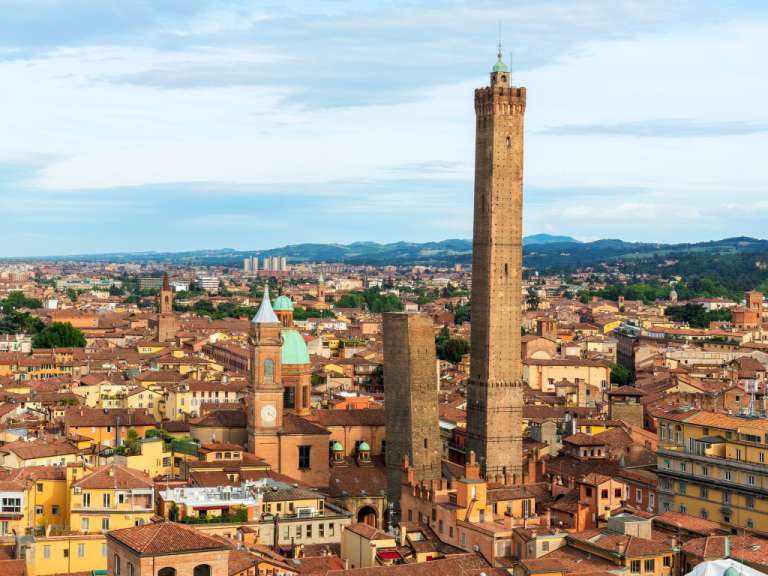 Das Bild zeigt eine Panoramaaufnahme der Stadt Bologna mit ihren charakteristischen Türmen und roten Dächern, gegen einen leicht bewölkten Himmel.