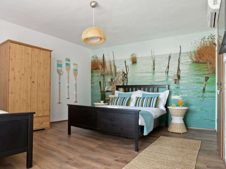 Ein Schlafzimmer mit einem dunklen Holzbett, einem holzfarbenen Kleiderschrank und einer Wanddekoration, die einen See mit Schilf und Stegen zeigt.