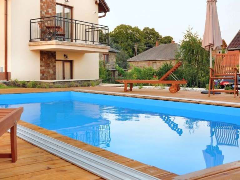 Ein Schwimmbecken mit blauem Wasser und einer Holzterrasse ist vor einem Haus mit einem Balkon zu sehen.