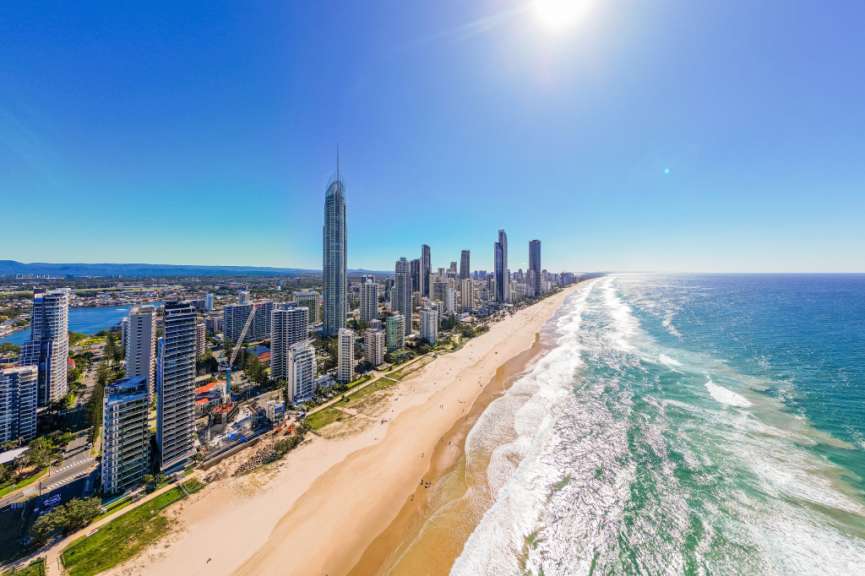 Ein Panorama zeigt eine breite Sandstrand, das Meer mit Wellen und eine Skyline von Hochhäusern unter einem blauen Himmel.