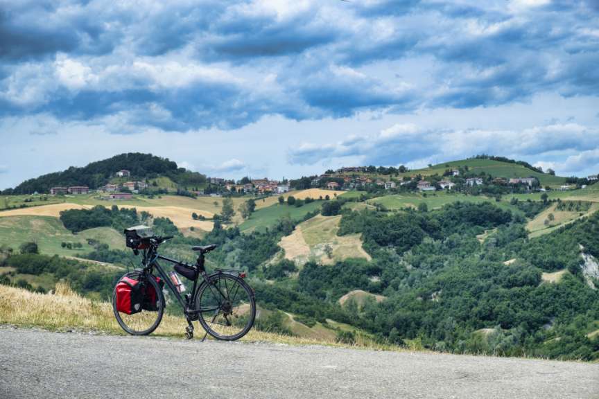 Ein beladenes Fahrrad steht am Straßenrand mit Blick auf eine hügelige Landschaft mit Wäldern, Feldern und Häusern unter einem bewölkten Himmel.