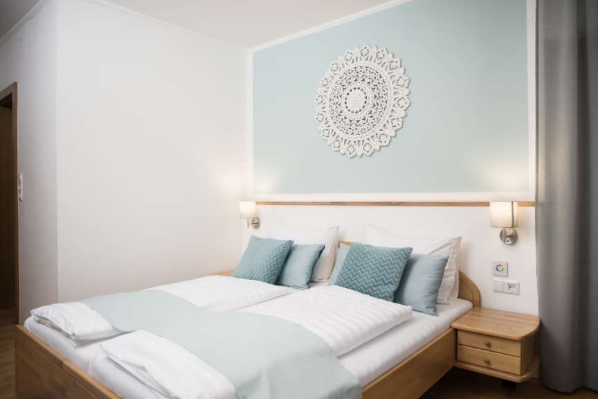 Ein Schlafzimmer mit einem Holzbett, zwei Kopfkissen, Bettwäsche in Weiß und Hellgrün sowie einer Wanddekoration und Wandlampen.
