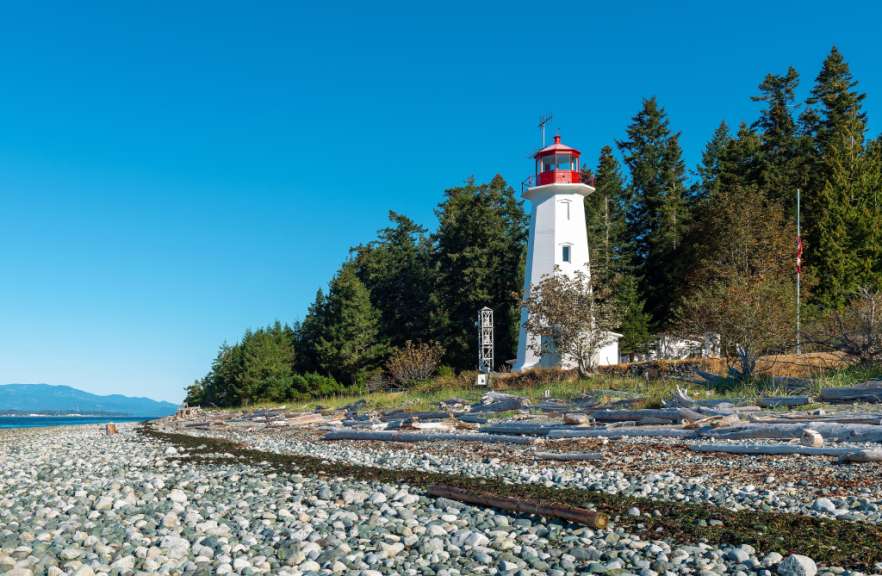Ein weißer Leuchtturm mit rotem Dach steht auf einer steinigen Küste, umgeben von Bäumen, mit blauem Himmel und Bergen im Hintergrund.