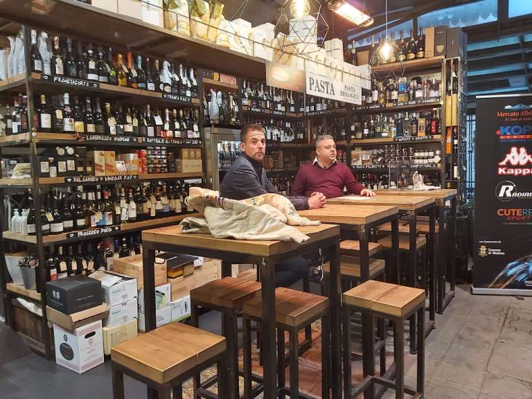 In einem Geschäft mit Wein, Lebensmitteln und verschiedenen Produkten sitzen zwei Männer an Holztischen.