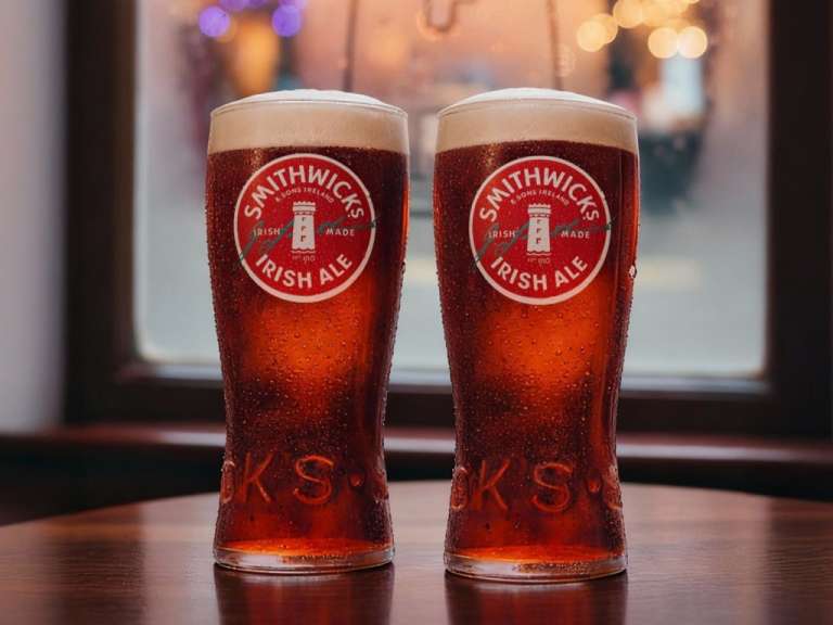 Zwei Gläser Smithwicks Irish Ale stehen auf einem Holztisch vor einem Fenster mit Lichtern im Hintergrund.